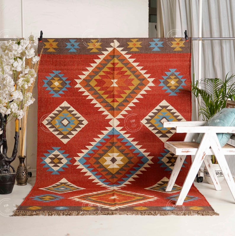 May include: A red rectangular rug with a geometric diamond pattern in shades of yellow, blue, and brown. The rug has a fringed edge and is displayed against a white background. The rug is a decorative floor covering.