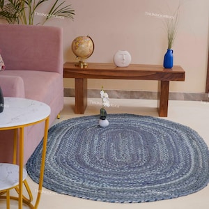 May include: An oval braided rug in shades of blue and grey, placed on a light floor. The rug is surrounded by furniture, including a pink sofa, a wooden bench, and two small side tables with yellow legs. Decorative items are placed on the furniture.