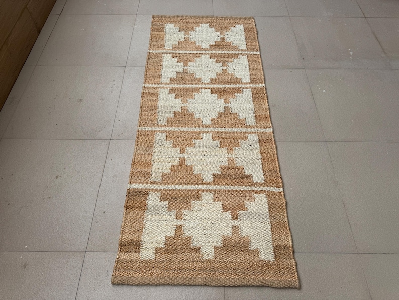 May include: A rectangular runner rug featuring a natural tan jute base with a repeating white geometric diamond pattern. The woven texture is visible throughout the surface, showcasing a series of five distinct diamond shapes aligned vertically.