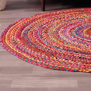 K&ouml;nnte beinhalten: Ein runder, geflochtener Teppich mit einer lebendigen Mischung aus Farben wie Rot, Rosa, Gelb und Blau. Der Teppich besteht aus gewebtem Stoff und hat ein kreisf&ouml;rmiges Muster. Er liegt auf einem hellen Holzboden.