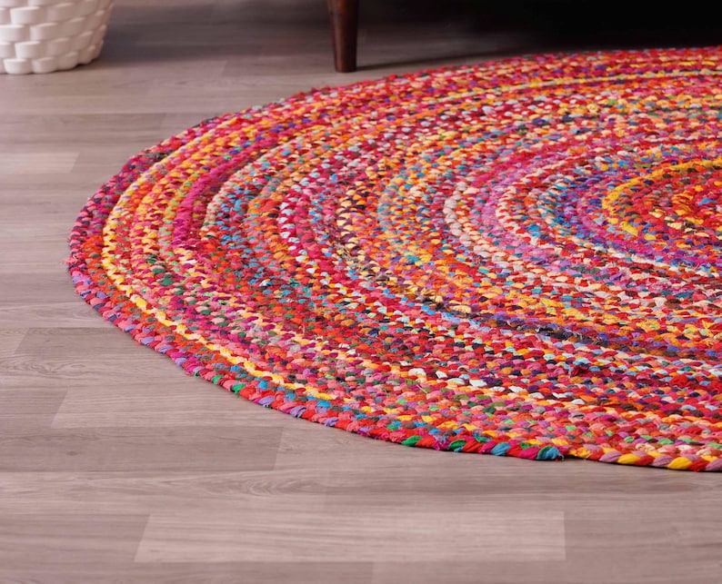 K&ouml;nnte beinhalten: Ein runder, geflochtener Teppich mit einer lebendigen Mischung aus Farben wie Rot, Rosa, Gelb und Blau. Der Teppich besteht aus gewebtem Stoff und hat ein kreisf&ouml;rmiges Muster. Er liegt auf einem hellen Holzboden.