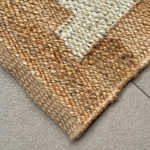 May include: A rectangular area rug woven from natural tan jute fibres featuring a distinct geometric pattern with a contrasting off-white section. The thick, braided texture of the plant-based material creates a durable surface with visible knots.