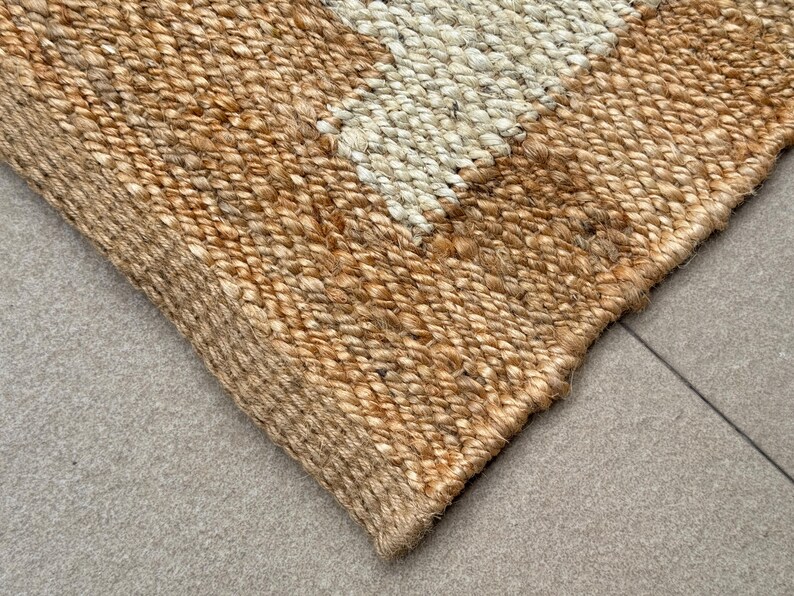 May include: A rectangular area rug woven from natural tan jute fibres featuring a distinct geometric pattern with a contrasting off-white section. The thick, braided texture of the plant-based material creates a durable surface with visible knots.