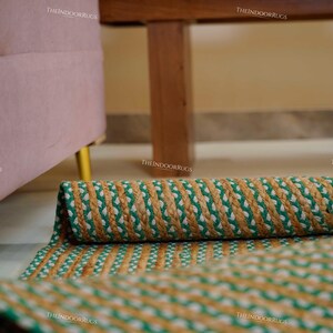 Puede incluir: Una alfombra de yute enrollada con un patr&oacute;n trenzado en verde, blanco y marr&oacute;n. La alfombra est&aacute; parcialmente desenrollada, mostrando el dise&ntilde;o detallado. Se encuentra sobre un suelo claro, con un sof&aacute; rosa y muebles de madera en el fondo.