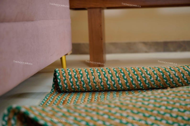 Puede incluir: Una alfombra de yute enrollada con un patr&oacute;n trenzado en verde, blanco y marr&oacute;n. La alfombra est&aacute; parcialmente desenrollada, mostrando el dise&ntilde;o detallado. Se encuentra sobre un suelo claro, con un sof&aacute; rosa y muebles de madera en el fondo.