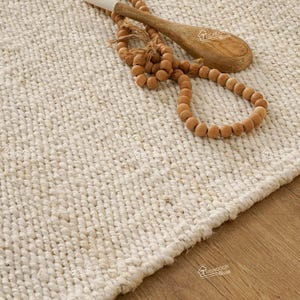 Könnte beinhalten: Ein weißer gewebter Teppich mit einem Holzlöffel und einer Schnur aus Holzperlen darauf.