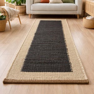 May include: A rectangular area rug with a dark gray center and a natural beige border. The rug is made of woven material and has a textured appearance. The rug is placed on a hardwood floor.