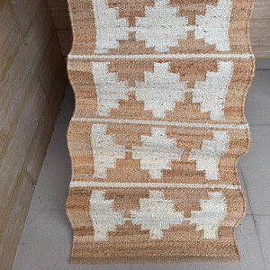 May include: A rectangular woven runner rug featuring a natural tan jute base with a repeating white geometric diamond pattern. The rug has a textured, fibrous surface and is displayed in a folded, cascading arrangement showing multiple sections.