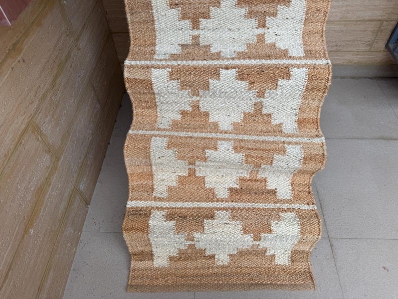 May include: A rectangular woven runner rug featuring a natural tan jute base with a repeating white geometric diamond pattern. The rug has a textured, fibrous surface and is displayed in a folded, cascading arrangement showing multiple sections.
