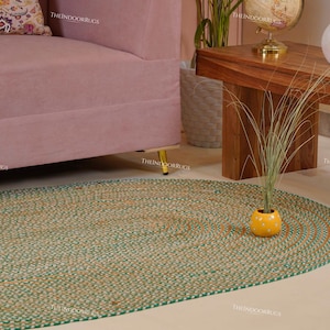 Puede incluir: Alfombra ovalada de yute con borde verde y tonos naturales. La alfombra est&aacute; colocada sobre un suelo de color claro, con un sof&aacute; rosa y una mesa de madera al fondo. Una peque&ntilde;a maceta amarilla con una planta est&aacute; sobre la alfombra.