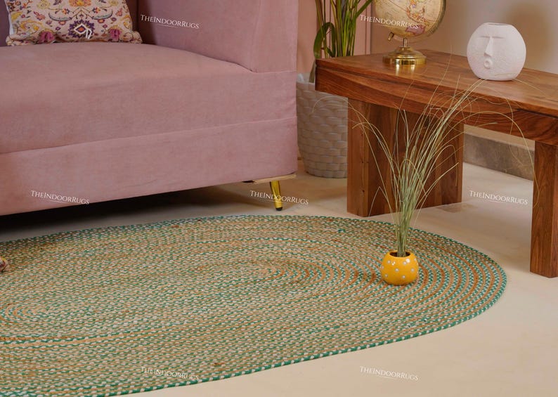 Puede incluir: Alfombra ovalada de yute con borde verde y tonos naturales. La alfombra est&aacute; colocada sobre un suelo de color claro, con un sof&aacute; rosa y una mesa de madera al fondo. Una peque&ntilde;a maceta amarilla con una planta est&aacute; sobre la alfombra.