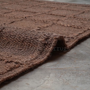 May include: A brown area rug with a textured, woven surface featuring a grid pattern of raised lines. The rug is folded at one corner, revealing the thick, fibrous material and the consistent brown color throughout the entire woven construction.