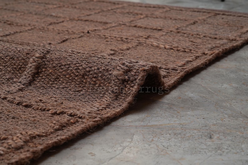 May include: A brown area rug with a textured, woven surface featuring a grid pattern of raised lines. The rug is folded at one corner, revealing the thick, fibrous material and the consistent brown color throughout the entire woven construction.