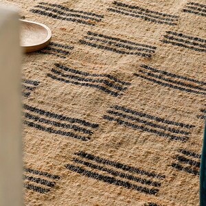 May include: Close-up of a woven rug with a natural beige background and repeating black horizontal stripe patterns. A small wooden bowl is visible in the upper left corner. The rug has a textured appearance.