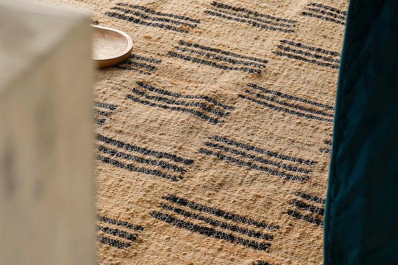 May include: Close-up of a woven rug with a natural beige background and repeating black horizontal stripe patterns. A small wooden bowl is visible in the upper left corner. The rug has a textured appearance.