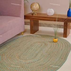 Puede incluir: Una alfombra ovalada de yute con tonos turquesa y naturales. La alfombra est&aacute; sobre un suelo claro, con un sof&aacute; rosa y una mesa de madera en el fondo. El dise&ntilde;o de la alfombra presenta patrones ovalados conc&eacute;ntricos.