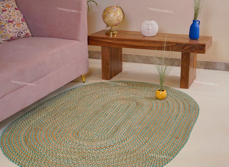 Puede incluir: Una alfombra ovalada de yute con tonos turquesa y naturales. La alfombra est&aacute; sobre un suelo claro, con un sof&aacute; rosa y una mesa de madera en el fondo. El dise&ntilde;o de la alfombra presenta patrones ovalados conc&eacute;ntricos.