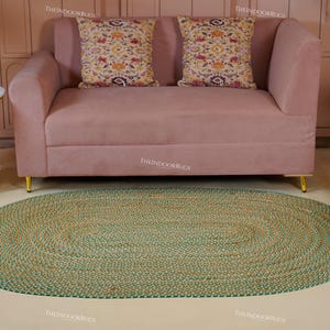 Puede incluir: Una alfombra ovalada de yute tejida a mano, con una combinaci&oacute;n de colores verde y natural. La alfombra est&aacute; colocada sobre un suelo claro, frente a un sof&aacute; rosa con cojines decorativos. No se especifican las dimensiones de la alfombra.