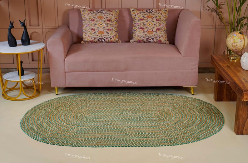 Puede incluir: Una alfombra ovalada de yute tejida a mano, con una combinaci&oacute;n de colores verde y natural. La alfombra est&aacute; colocada sobre un suelo claro, frente a un sof&aacute; rosa con cojines decorativos. No se especifican las dimensiones de la alfombra.