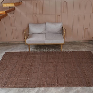 May include: A rectangular brown area rug featuring a grid pattern of raised square sections. The rug is positioned on a light gray floor in front of a light gray two-seater sofa. The text theindoorrugs is visible in the center of the rug.