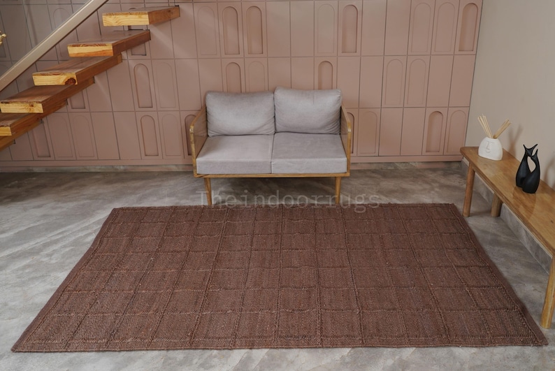 May include: A rectangular brown area rug featuring a grid pattern of raised square sections. The rug is positioned on a light gray floor in front of a light gray two-seater sofa. The text theindoorrugs is visible in the center of the rug.