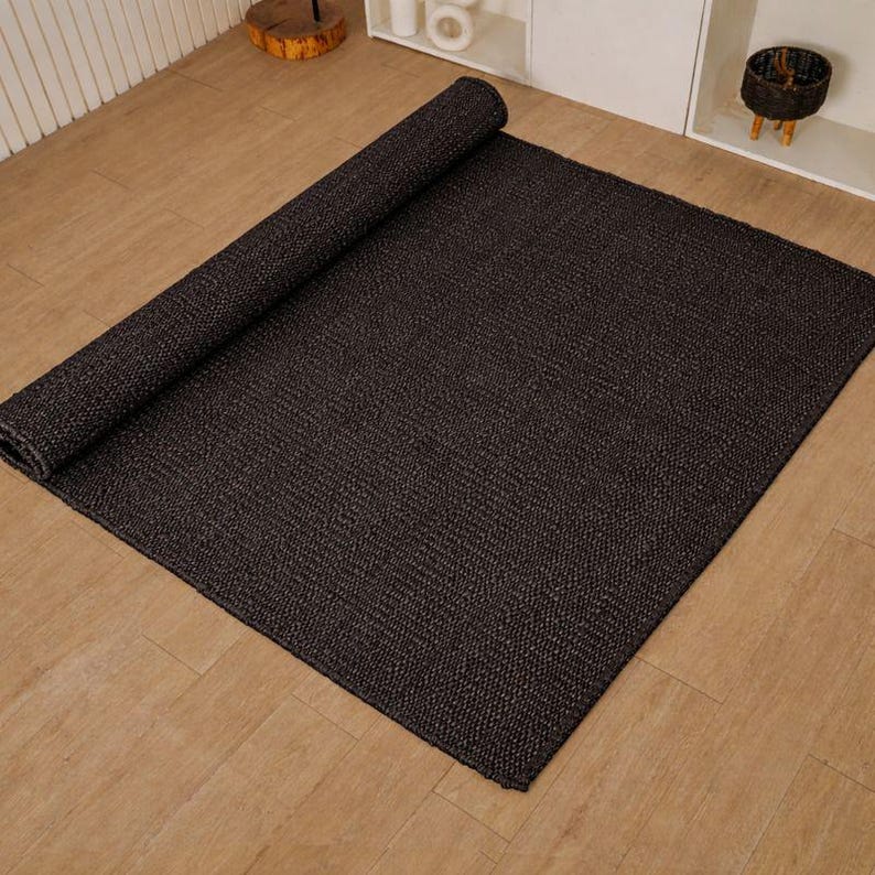 May include: A dark brown rectangular rug, partly rolled, on a light brown wooden floor. The rug has a woven texture and a solid colour. The rolled section is at the top left. The rug is suitable for a living room.