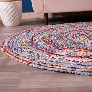 K&ouml;nnte beinhalten: Ein runder, geflochtener Teppich mit einem Spiraldesign. Der Teppich hat eine Mischung aus Farben, darunter Blau, Rot, Gelb und Orange. Der Teppich besteht aus Stoff und liegt auf einem Holzboden.