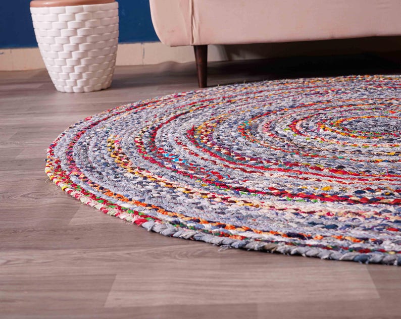 K&ouml;nnte beinhalten: Ein runder, geflochtener Teppich mit einem Spiraldesign. Der Teppich hat eine Mischung aus Farben, darunter Blau, Rot, Gelb und Orange. Der Teppich besteht aus Stoff und liegt auf einem Holzboden.