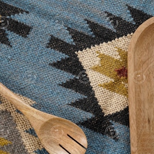 May include: A wooden slotted spoon with a white handle rests on a blue, black, and beige patterned rug. A wooden serving tray is positioned to the right of the spoon and rug.