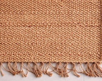 May include: Close-up of a woven jute rug with a natural tan color. The rug has a textured surface with a diagonal weave pattern. The bottom edge features a fringe detail made of the same material.