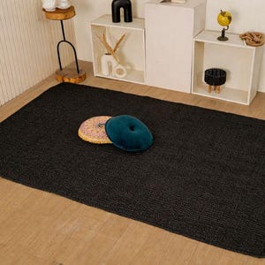 May include: A dark grey rectangular rug with a textured weave. Two decorative cushions, one teal and one multi-coloured, are placed on the rug. The rug is on a light wooden floor, with shelves and decor in the background.