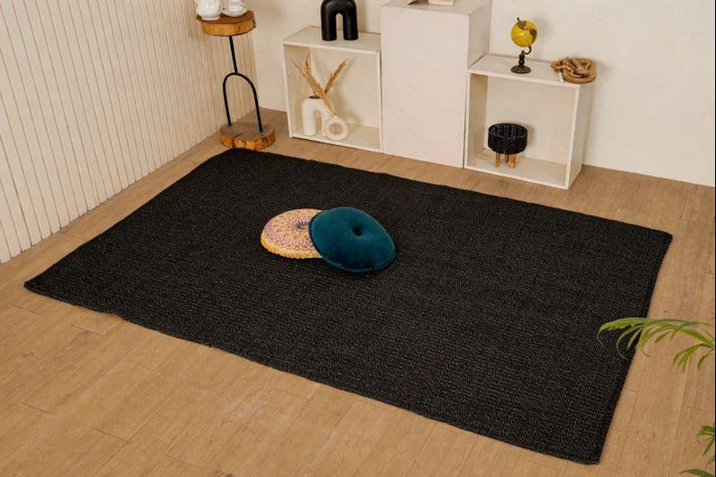 May include: A dark grey rectangular rug with a textured weave. Two decorative cushions, one teal and one multi-coloured, are placed on the rug. The rug is on a light wooden floor, with shelves and decor in the background.