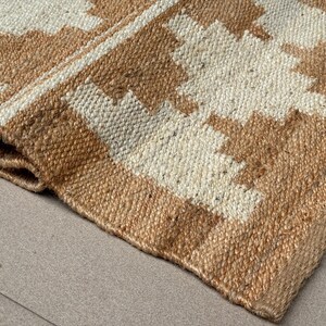 May include: A woven rug featuring a geometric pattern in natural tan and off-white fibres. The material has a coarse, textured appearance with a visible braided weave. One corner of the rug is folded over, revealing the thickness of the material.