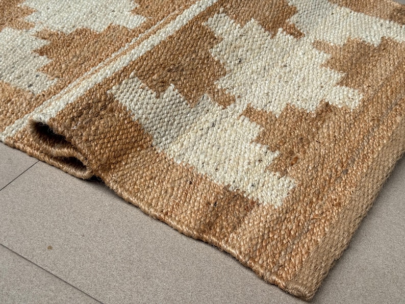 May include: A woven rug featuring a geometric pattern in natural tan and off-white fibres. The material has a coarse, textured appearance with a visible braided weave. One corner of the rug is folded over, revealing the thickness of the material.