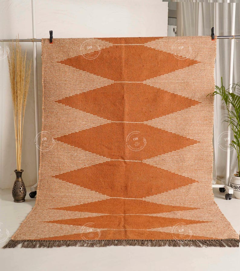 May include: A large, rectangular rug with a geometric diamond pattern in shades of orange and beige. The rug has a fringed edge and is displayed against a neutral background. The design features alternating rows of diamonds and horizontal stripes.