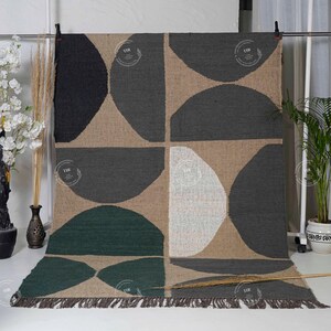May include: A large, rectangular rug with a geometric pattern. The design features large, semi-circular shapes in black, gray, green, and white against a tan background. The rug has a fringed edge and is displayed against a white backdrop.