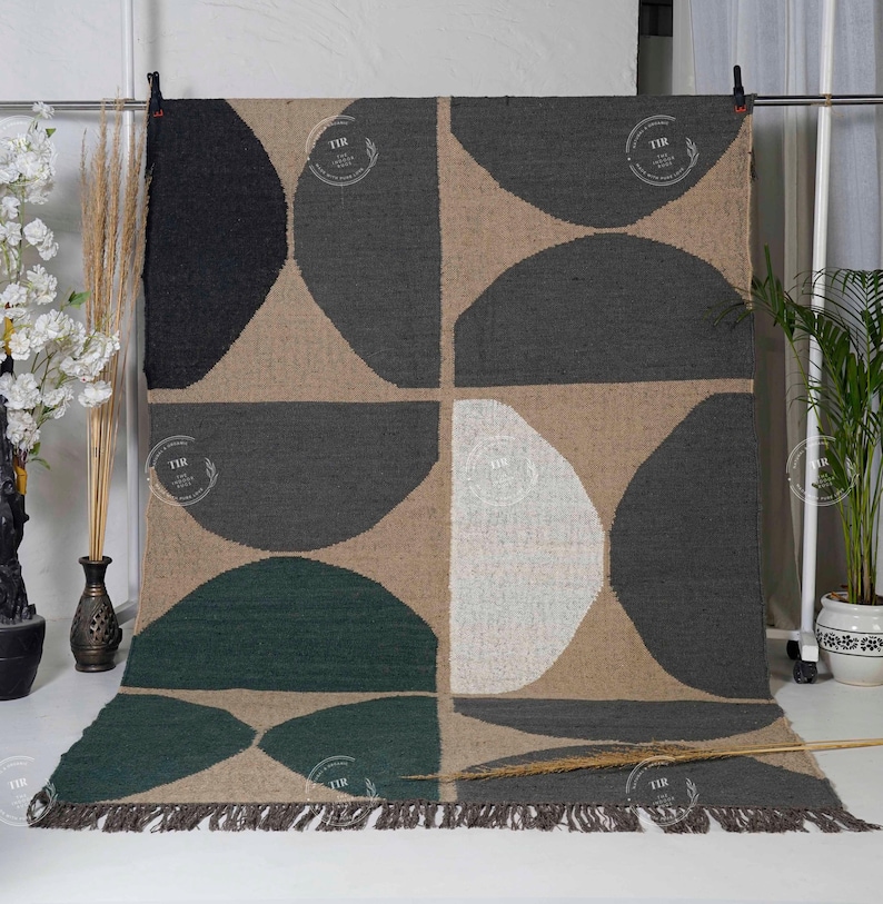 May include: A large, rectangular rug with a geometric pattern. The design features large, semi-circular shapes in black, gray, green, and white against a tan background. The rug has a fringed edge and is displayed against a white backdrop.