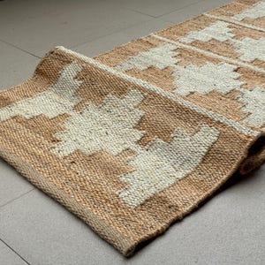 May include: A rectangular runner rug featuring a woven texture with a natural tan base colour and a repeating geometric pattern in off-white. The rug has a thick, fibrous construction with a visible braided weave and a curled edge at one end.