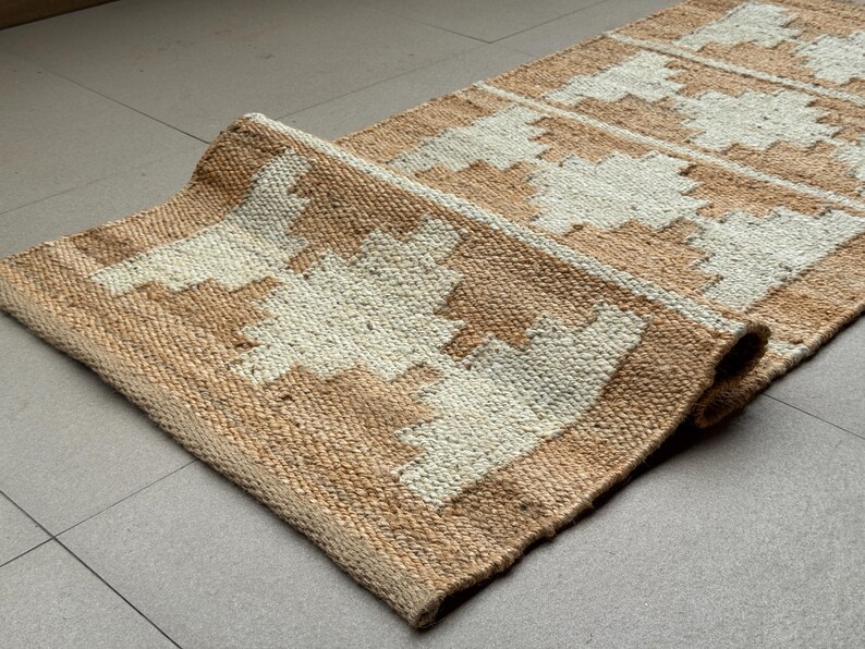 May include: A rectangular runner rug featuring a woven texture with a natural tan base colour and a repeating geometric pattern in off-white. The rug has a thick, fibrous construction with a visible braided weave and a curled edge at one end.
