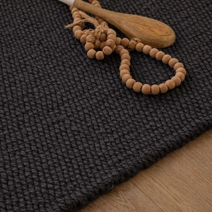 May include: Close-up of a dark grey woven rug with a textured surface. A wooden spoon and a strand of wooden beads rest on the rug. The rug is placed on a light brown wooden floor.