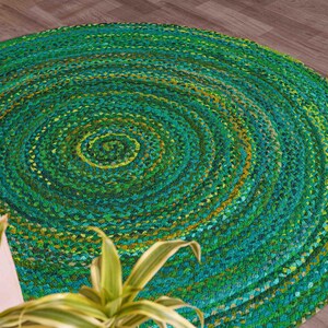 Puede incluir: Una alfombra redonda trenzada en tonos verdes y amarillos, creando un patrón en espiral. La alfombra está hecha de un material tejido y está colocada sobre un suelo de madera. El diseño de la alfombra añade un elemento natural y texturizado a la habitación.