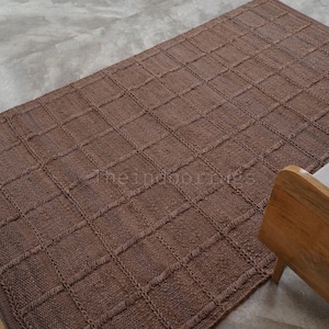 May include: A rectangular brown area rug featuring a raised grid pattern created by textured, braided-style lines. The rug has a consistent solid brown color throughout and displays the text theindoorgrues in the center of the woven surface.