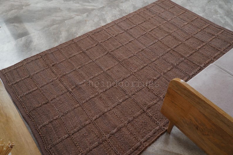 May include: A rectangular brown area rug featuring a raised grid pattern created by textured, braided-style lines. The rug has a consistent solid brown color throughout and displays the text theindoorgrues in the center of the woven surface.