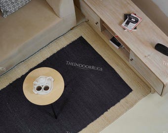 May include: Overhead view of a living room setup featuring a black and beige rug with the text "THEINDOORRUGS". A small, round wooden table holds a white teapot and cups. A light-colored sofa and wooden cabinet are also visible.