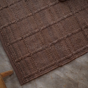 May include: A brown knitted rug featuring a raised grid pattern of squares. The textile has a consistent textured surface with a thick border edge. The design consists of vertical and horizontal lines forming a repeating square grid across the fabric.