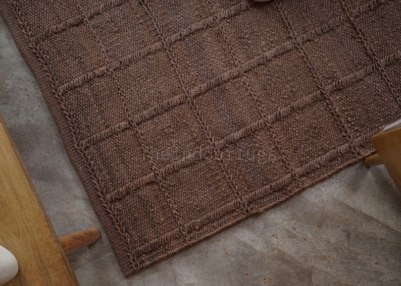 May include: A brown knitted rug featuring a raised grid pattern of squares. The textile has a consistent textured surface with a thick border edge. The design consists of vertical and horizontal lines forming a repeating square grid across the fabric.