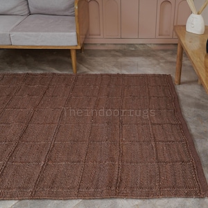 May include: A rectangular brown area rug featuring a grid pattern of raised, textured lines. The rug has a woven appearance and includes the text "theindoorrugs" printed in a light color across the center of the surface, showcasing a consistent weave.