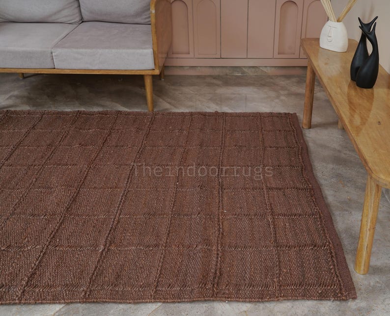 May include: A rectangular brown area rug featuring a grid pattern of raised, textured lines. The rug has a woven appearance and includes the text "theindoorrugs" printed in a light color across the center of the surface, showcasing a consistent weave.
