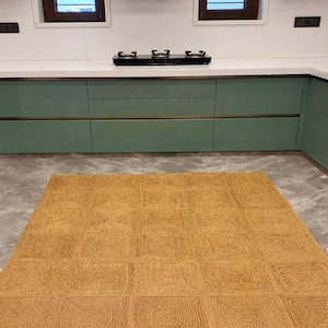 May include: A kitchen interior featuring a woven jute rug with a geometric pattern. The rug is placed on a gray tiled floor in front of green cabinets and a white countertop with a black stovetop.