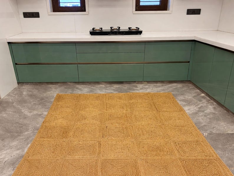 May include: A kitchen interior featuring a woven jute rug with a geometric pattern. The rug is placed on a gray tiled floor in front of green cabinets and a white countertop with a black stovetop.